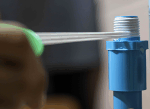 Hand applying teflon tape to blue plumbing pipe, wrapping at the base of the threads (closest to the pipe body).